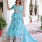 "Full-length view of turquoise lehenga set with peplum blouse, tiered skirt with multi-colored embroidered border, flowing dupatta, and pearl jewelry on colonial architecture background"
