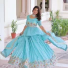 "Woman in turquoise lehenga with peplum blouse positioned against white marble column with green landscape, wearing coordinating dupatta and traditional jewelry for festive occasion"